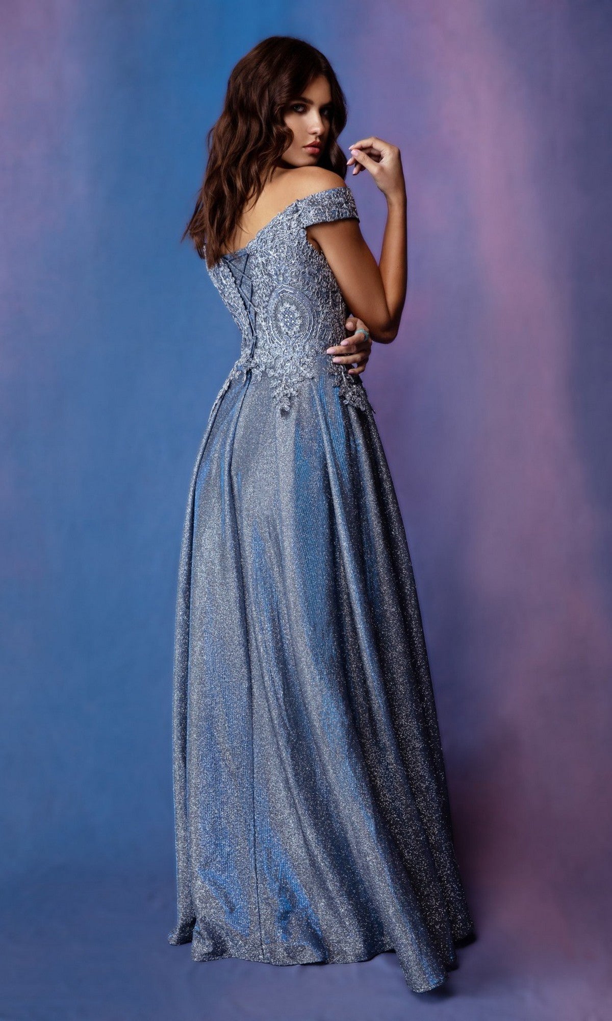 A woman in the Long Prom Dress: Eureka 9806—a sparkling blue, off-the-shoulder, floor-length gown with a glitter a-line skirt—stands against a soft purple and blue gradient background, facing away but looking back over her shoulder.